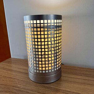 May include: A silver metal lamp with a square grid pattern on the shade. The lamp is turned on and the light shines through the grid.