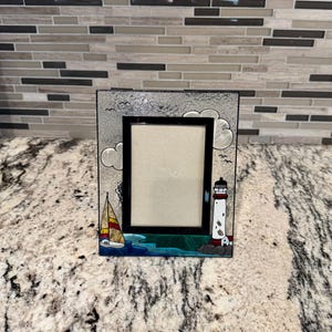May include: A rectangular picture frame with a nautical theme. The frame features a black border and a glass front. The design includes a sailboat, a lighthouse, clouds, and birds. The frame is painted with blue, white, red, and yellow accents.