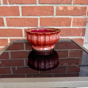 Puede incluir: Un cuenco de cerámica roja con un diseño acanalado y un acabado brillante. El cuenco se encuentra sobre una superficie reflectante, con una pared de ladrillos al fondo. El cuenco es probablemente para fines decorativos.