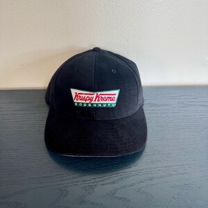 May include: A black baseball cap featuring an embroidered patch on the front. The patch displays the red text Krispy Kreme above the green text DOUGHNUTS, all enclosed within a white trapezoidal border with a green outline.