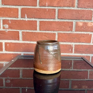 May include: A handmade ceramic jar with a dark brown glaze and lighter brown accents. The jar has a cylindrical shape with a slightly flared rim and a rough, textured surface. The jar is sitting on a reflective surface.