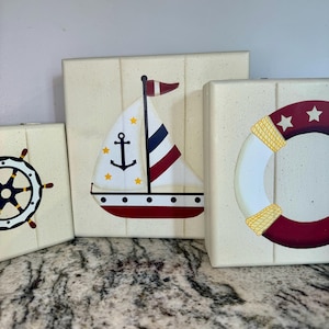 May include: Three wooden blocks with nautical themed designs. The first block features a red, white and blue sailboat with a yellow anchor and stars. The second block has a red and white life preserver with a yellow rope and white stars. The third block has a black and white ship's wheel.