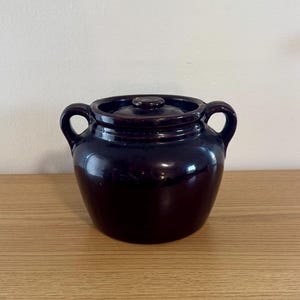 May include: A dark brown ceramic bean pot with a matching lid and two side handles. The pot features a smooth, glossy glaze and a rounded body shape, providing a classic look for kitchen storage or cooking applications.