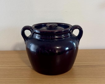 Può includere: Un vaso per fagioli in ceramica marrone scuro con coperchio abbinato e due manici laterali. Il vaso presenta una smaltatura liscia e lucida e una forma arrotondata, offrendo un aspetto classico per la conservazione in cucina.