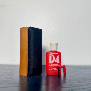 May include: A record cleaning kit featuring a wooden-handled brush with black fabric, a red plastic bottle of D4+ Hi-Technology Record Cleaning Fluid with a white cap, and a small red plastic brush with black bristles for stylus cleaning.