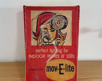 Op de afbeelding: Een rechthoekige rode kartonnen doos voor een ACME-LITE mov-E-lite cameralamp. De voorkant bevat een afbeelding van een persoon die een camera met een lamp vasthoudt, met de tekst: perfect lighting for INDOOR movies or stills, ACME-LITE, mov-E-lite.