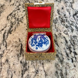 May include: A small, round, white and blue porcelain trinket box with a dragon design, inside a decorative box with a red satin interior. The outer box has a gold and brown patterned design.