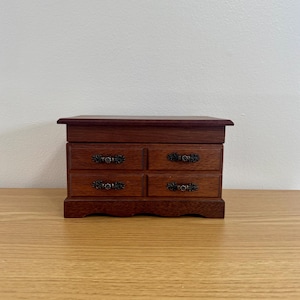 May include: A small, wooden jewelry box with four drawers. The box is a dark reddish-brown color with ornate, dark metal drawer pulls. The box sits on a light-colored wooden surface against a plain white background.