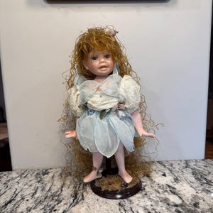Puede incluir: Muñeca de porcelana con pelo largo y rizado de color rojizo y ojos azules. La muñeca lleva un vestido azul claro y blanco con detalles en forma de pétalos. Se apoya sobre una base redonda de color marrón oscuro con textura de musgo, y es un objeto de colección decorativo.
