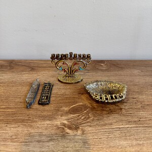 Puede incluir: Una colección de objetos religiosos judíos sobre una superficie de madera. Incluye una menorá de latón con detalles coloridos, dos pequeñas mezuzás y un plato pequeño con la palabra "Shalom" inscrita.