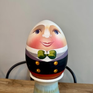 May include: A hand-painted egg with a whimsical face, wearing a black suit with a green bow tie and gold buttons. The egg is sitting on a white ceramic stand.