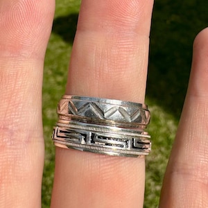 May include: Two silver rings stacked on a finger. The top ring has a textured design with a series of triangles. The bottom ring has a black and silver Greek key design.
