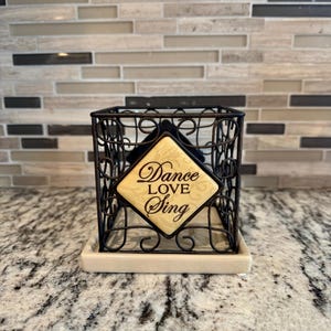 May include: A black metal wire organiser with a decorative scroll design. It has a beige ceramic base and a square, yellow sign that reads "Dance Love Sing".