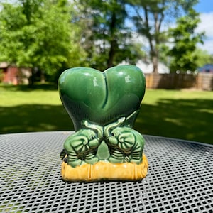 Op de afbeelding: Een groene keramische olifantenfiguur met twee tegenover elkaar staande olifanten, op een gele basis. De figuur heeft een hartvormige bovenkant en staat buiten op een metalen tafel.