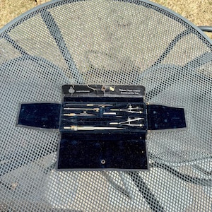 Pode incluir: Um estojo de couro preto vintage com interior de veludo, contendo um conjunto de ferramentas de desenho. As ferramentas são de latão e incluem um compasso, um transferidor e uma régua. O estojo é etiquetado como "Technical Supply Company, Inc.  Exclusive New York"