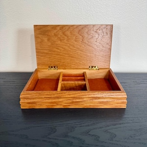 May include: Open wooden jewelry box with a hinged lid. The box is made of light brown wood and has a rectangular shape. The interior is lined with a reddish-brown fabric and divided into several compartments. The box is sitting on a black surface.