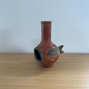 Puede incluir: Un jarrón de cerámica hecho a mano con forma de pez. El jarrón es principalmente de color marrón rojizo con detalles verdes y azules en las aletas y la boca. Tiene un cuello cilíndrico y una amplia abertura para exhibir.
