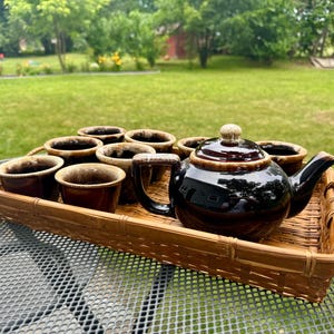 May include: A brown ceramic tea set with a teapot and several cups, presented on a woven bamboo tray. The teapot is dark brown and glossy, with a light-colored knob on the lid. The cups are a lighter brown color.