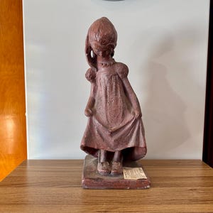 Op de afbeelding: Een terracotta beeldje van een meisje in een jurk, dat de zoom van haar rok vasthoudt. Het beeldje staat op een rechthoekige basis.