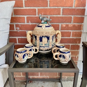 Könnte beinhalten: Ein Set aus sechs Keramik-Bierkrügen mit blauen und weißen Designs. Die Krüge sind um einen großen Keramik-Bierkrug mit einer Hahnfigur auf der Oberseite angeordnet. Die Krüge stehen auf einem Tisch mit einer schwarzen Glasplatte und einem Metallrahmen.