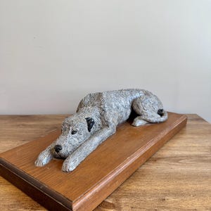 Può includere: Una statuetta grigia scolpita di un lupo irlandese sdraiato su una base di legno. La statuetta è dettagliata con la pelliccia e ha gli occhi neri.