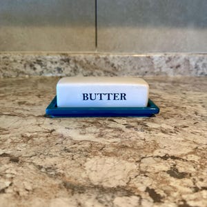 May include: A rectangular ceramic butter dish featuring a white lid with the word BUTTER printed in dark blue letters, resting on a solid dark blue base. The dish has a smooth, glossy finish and a clean, simple design for kitchen storage.