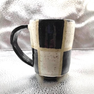 Puede incluir: Una taza de cerámica negra y blanca con un patrón a cuadros. La taza tiene un asa y está salpicada de esmalte marrón.