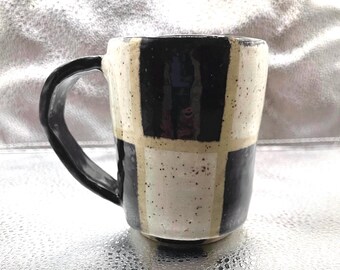 Puede incluir: Una taza de cerámica negra y blanca con un patrón a cuadros. La taza tiene un asa y está salpicada de esmalte marrón.