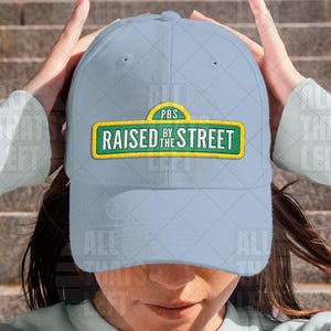 May include: A light blue baseball cap with the text "RAISED BY THE STREET" in a green and yellow design. The cap has a curved brim and a classic six-panel construction. The hat is made of a soft material.