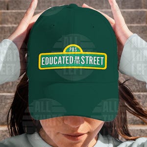 May include: A dark green baseball cap with the text "EDUCATED ON THE STREET" in yellow and white. The cap has a curved brim and a PBS logo. The hat is being held up by a person.