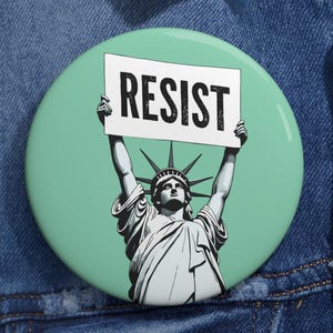 May include: A green button with a white background and the word "RESIST" in black text. The Statue of Liberty is holding the sign in front of her.