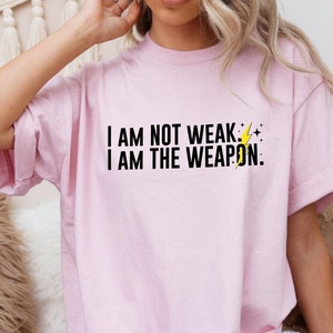 Puede incluir: Una camiseta rosa con el texto "I AM NOT WEAK. I AM THE WEAPON." en letras negras y un gr&aacute;fico de rayo amarillo.