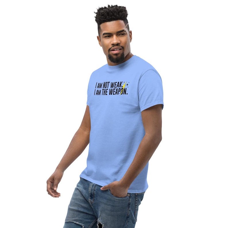 Puede incluir: Una camiseta azul claro con un gr&aacute;fico negro que dice "I AM NOT WEAK. I AM THE WEAPON." con un s&iacute;mbolo de rayo amarillo.