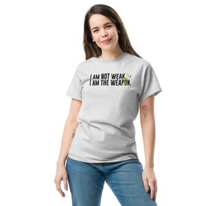 Puede incluir: Una camiseta gris con el texto "I AM NOT WEAK. I AM THE WEAPON." en letras negras. Un gr&aacute;fico de rayo amarillo est&aacute; al lado del texto.