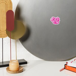 May include: A pink sticker with the text "Fuck ICE" in a stylized font. The sticker is on a gray surface. The image also includes a decorative metal sculpture, a candle in a jar, and a stack of books.