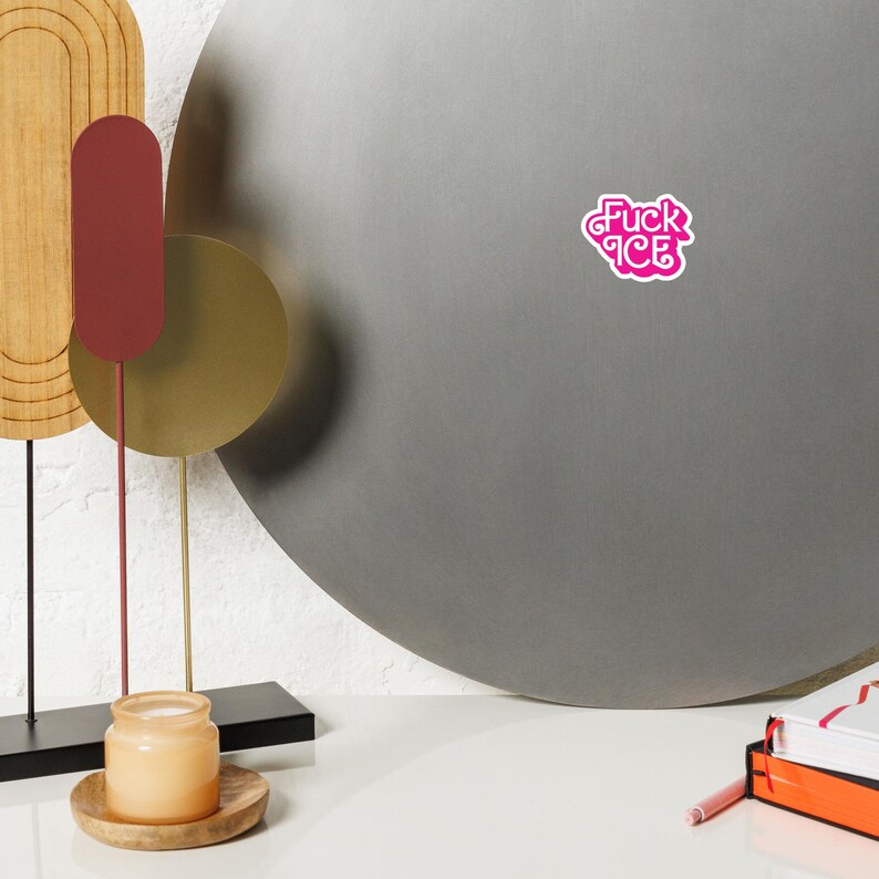 May include: A pink sticker with the text "Fuck ICE" in a stylized font. The sticker is on a gray surface. The image also includes a decorative metal sculpture, a candle in a jar, and a stack of books.