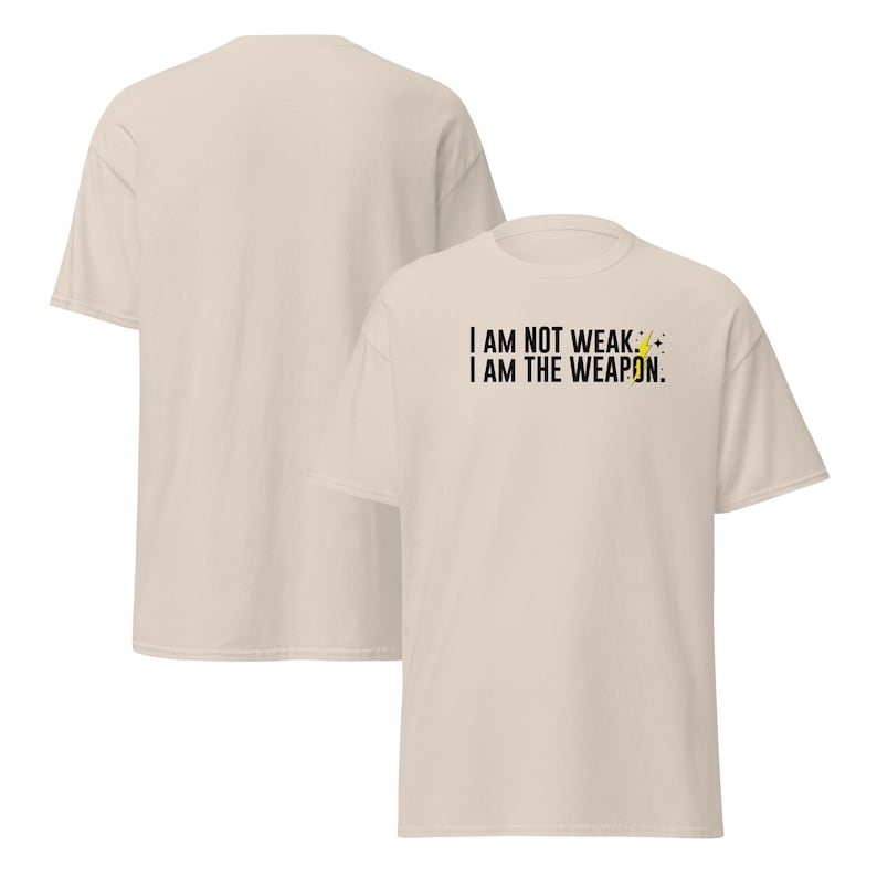 Puede incluir: Una camiseta beige con el texto "I AM NOT WEAK. I AM THE WEAPON." impreso en la parte delantera. Un rayo gr&aacute;fico est&aacute; al lado del texto.