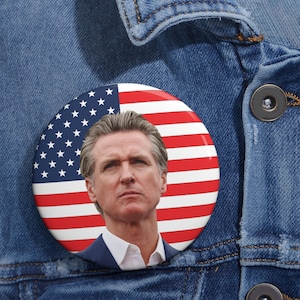 May include: A round button featuring the American flag design with a portrait of a man in a suit. The button is pinned to a blue denim jacket.