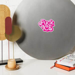 May include: A pink sticker with the words "Fuck ICE" in a stylized font. The sticker is on a gray, circular surface. The image also includes decorative objects and books.