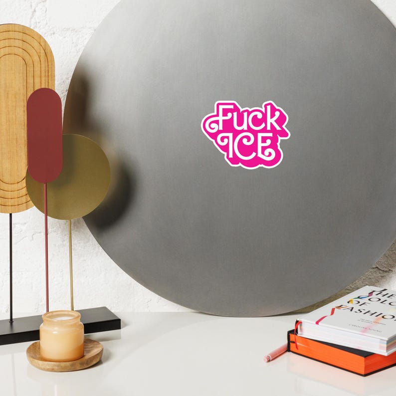 May include: A pink sticker with the words "Fuck ICE" in a stylized font. The sticker is on a gray, circular surface. The image also includes decorative objects and books.