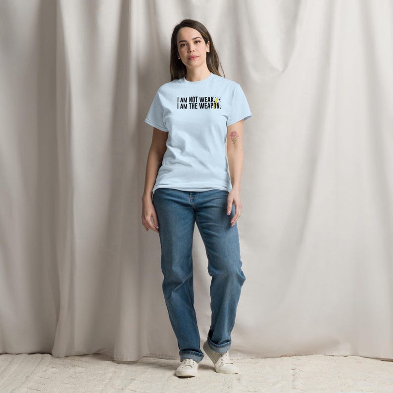 Puede incluir: Una camiseta azul claro con el texto "I AM NOT WEAK, I AM THE WEAPON." impreso en negro. La persona que lleva la camiseta est&aacute; de pie frente a un fondo blanco.