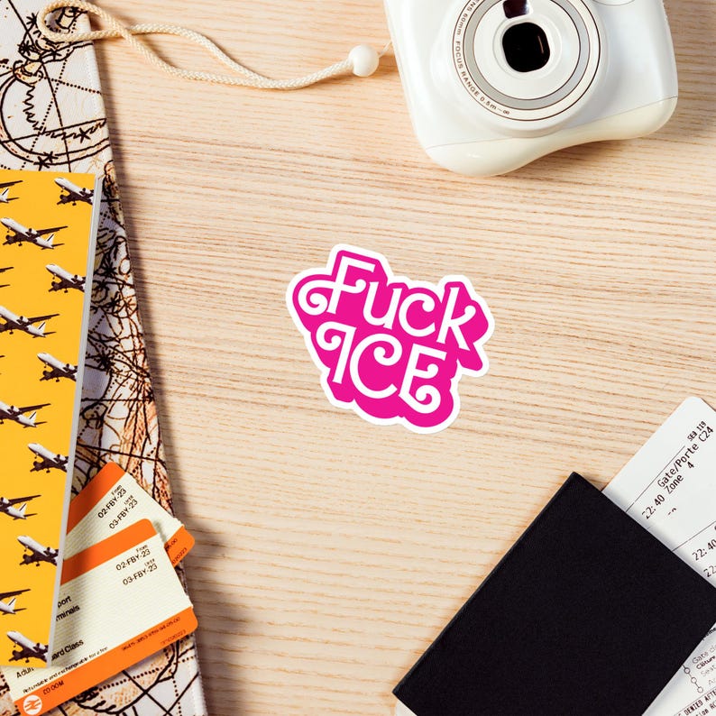May include: A pink sticker with the words "Fuck ICE" in a stylized font. The sticker is on a wooden surface, surrounded by travel-related items like a camera, passport, and boarding passes.
