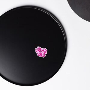 May include: A pink sticker with the words "Fuck ICE" in a stylized font. The sticker is on a black surface, with a portion of another black object visible in the background.