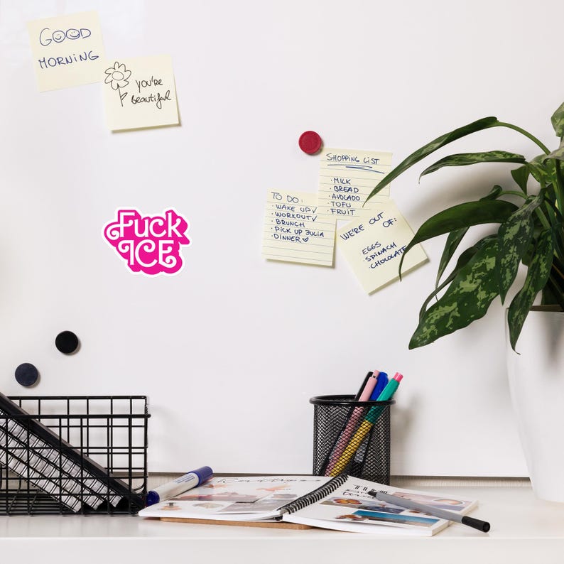 May include: A pink sticker with the words "Fuck Ice" in a stylized font is affixed to a white surface. Other sticky notes with handwritten text are also visible, along with a shopping list and a to-do list. A potted plant and office supplies are in the background.