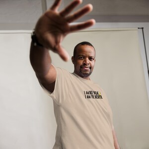 Puede incluir: Una persona con una camiseta beige con el texto "I AM NOT WEAK. I AM THE WEAPON." impreso en negro.