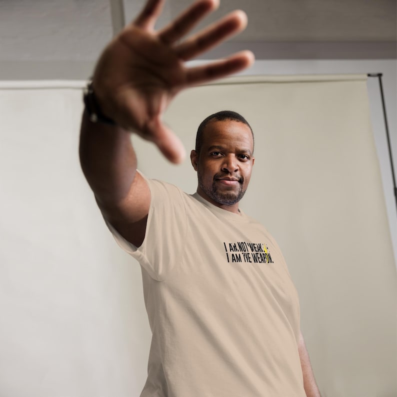 Puede incluir: Una persona con una camiseta beige con el texto "I AM NOT WEAK. I AM THE WEAPON." impreso en negro.