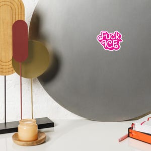 May include: A pink sticker with the words "Fuck ICE" in a stylized font. The sticker is on a gray surface. The image also includes a decorative metal sculpture, a candle in a jar, and a stack of books.