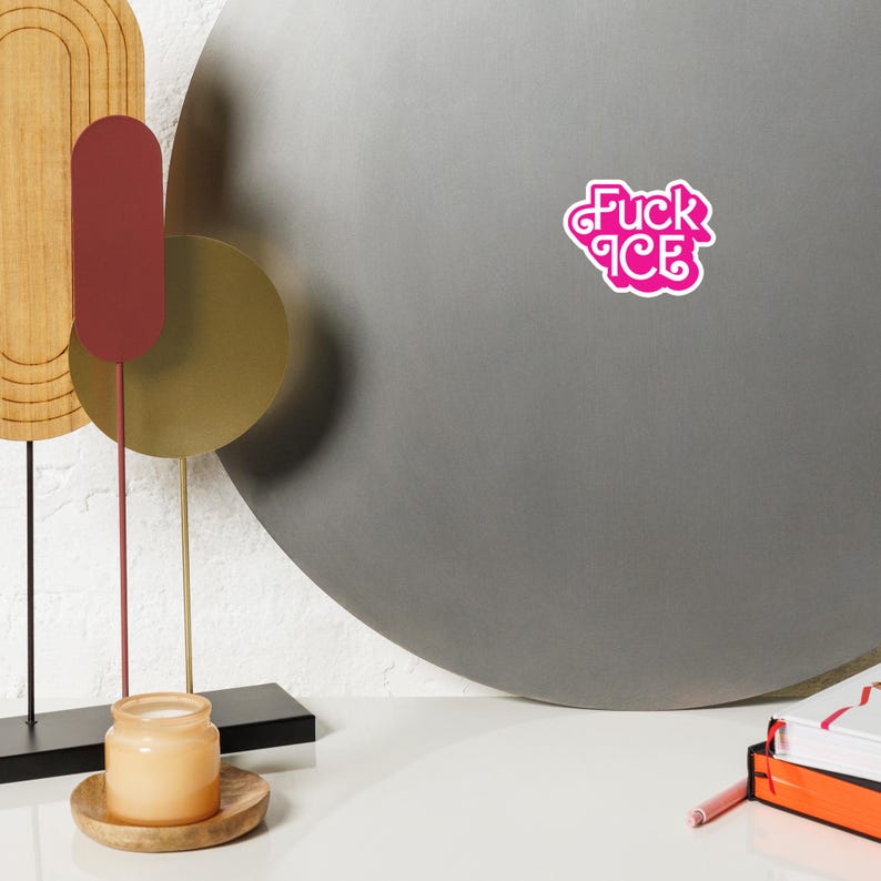 May include: A pink sticker with the words "Fuck ICE" in a stylized font. The sticker is on a gray surface. The image also includes a decorative metal sculpture, a candle in a jar, and a stack of books.