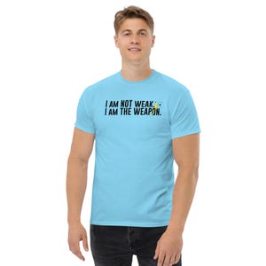 Puede incluir: Una camiseta azul claro con el texto "I AM NOT WEAK. I AM THE WEAPON." en letras negras y un gr&aacute;fico de rayo amarillo.
