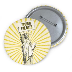 May include: A yellow and white button with a vintage-style illustration of the Statue of Liberty holding a sign that says "Uphold the Oath".